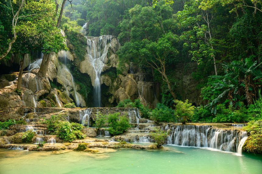 Golden sunlight illuminates the layered limestone cascades and emerald pools of Kuang Si Waterfalls in northern Laos. Dense tropical forest frames the flowing water, highlighting the vibrant greenery and natural textures.