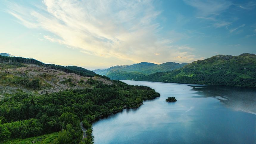 Loch Lomond, Scottish highlands, a tranquil lake scene under a bright blue sky with fluffy white clouds. Lush green hills adorned with dense trees border the water, creating a serene landscape