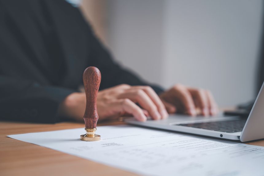 Wooden stamp on an approval document with a professional person typing on a laptop symbolizing authorization validation office workflow legal process paperwork and business administration