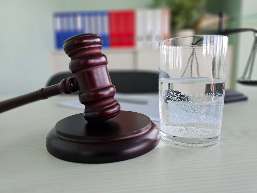 A gavel sits on a wooden base next to a glass of water. Documents and office supplies are visible in the background, indicating a legal office environment.