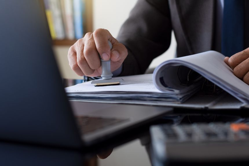 Businessman in suit hand stamping rubber stamp on document in file folder with laptop computer on the desk at office. Authorized allowance permission approval concept.