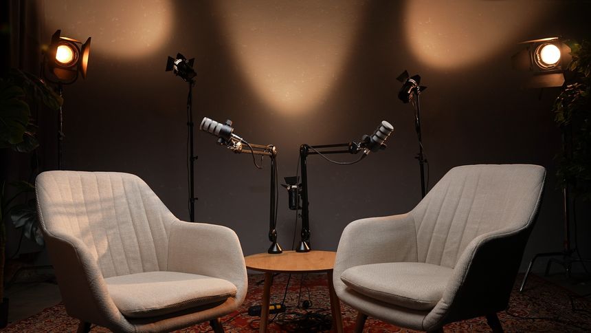 In a thoughtfully designed podcast studio, two stylish chairs face each other across a table, with microphones poised for an engaging interview. Soft light highlights the inviting space.