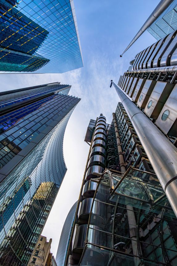 Low angle upward exterior view of the Lloyd Building, an icon of high-tech architecture with other skyscrapers with concave blue glass facades reflecting the sky and buildings under a clear blue sky.