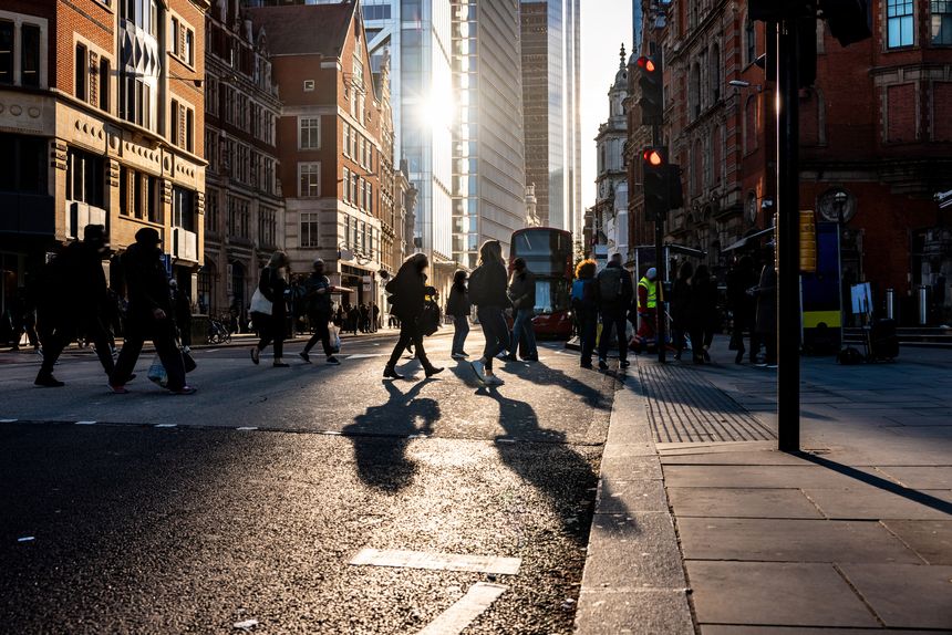 Backlit commuters and tourists walking on a crosswalk in central London during sunset, casting long shadows across the asphalt while surrounded by urban architecture and skyscrapers