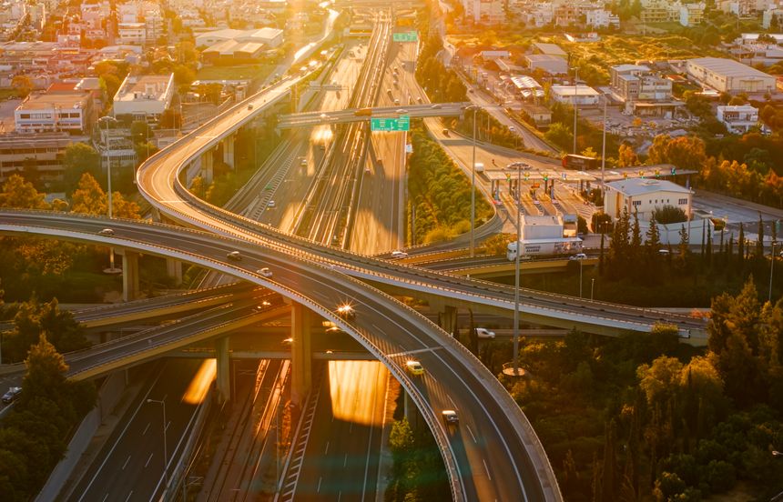 Multiple elevated expressways and an intricate highway interchange connect busy urban roads with moving vehicles during a golden hour sunset, showing modern transportation infrastructure