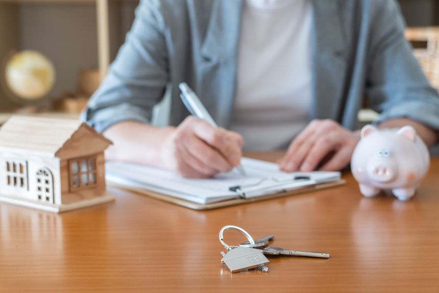 Person signing mortgage contract for a new home purchase on desk with house model, keys and piggy bank, symbolizing real estate financing, savings and property investment planning