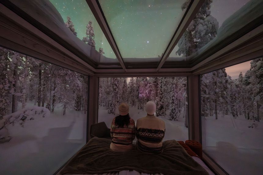 Relaxing in a unique glass-roofed cabin, two travelers gaze at the mesmerizing Northern Lights illuminating the snow-covered landscape of Akaslompolo, Finland.