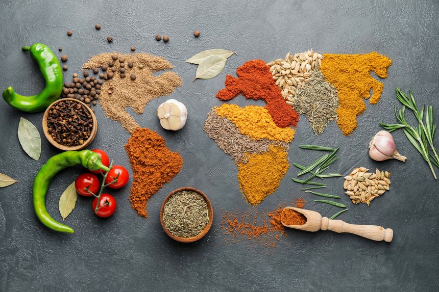 This vibrant, high-angle photograph symbolizes the global journey and cultural diversity of flavors through food. The continents are rendered using different types of ground spices?curry, paprika, turmeric, cumin, and seeds?arranged on a slate gray texture. Fresh ingredients like green chili peppers, cloves, garlic, and rosemary sprigs surround the spice map. Excellent for food blogs, international cuisine advertisements, or culinary travel magazines.
