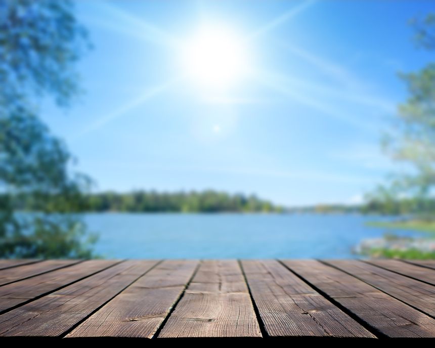 Empty wooden terrace overlooking a lake under a bright sunny sky. The blurred natural background with trees, blue water, and lens flare creates a peaceful summer atmosphere. Stock photo.