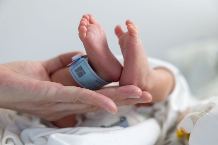 A baby's feet are held up by a person. The baby is wearing a blue tag. The baby is in a hospital bed