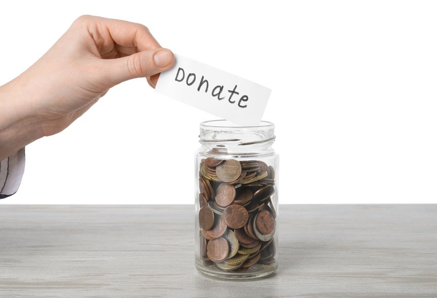 Woman holding paper note with word Donate near jar full of coins at wooden table against white background, closeup