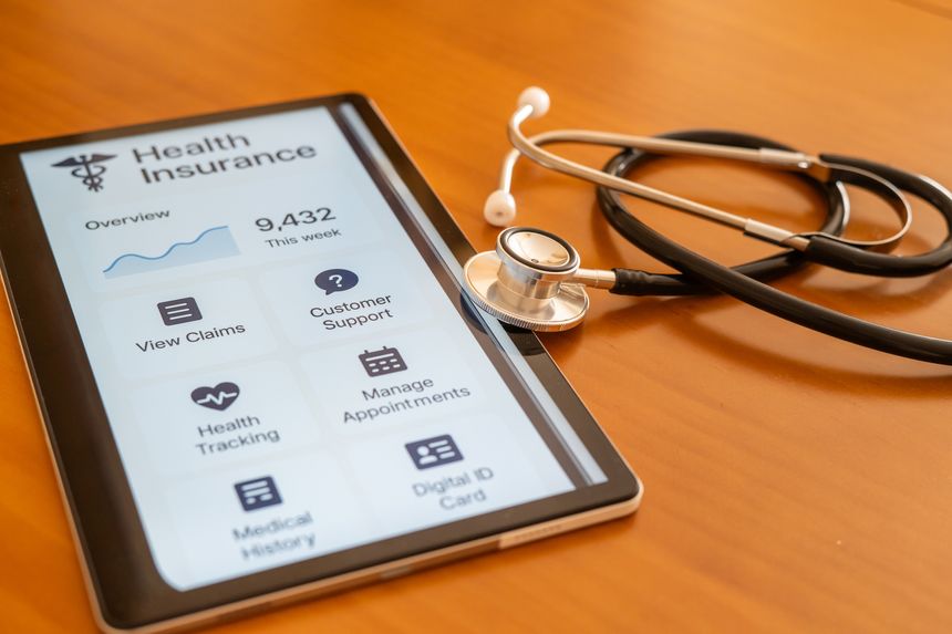 Tablet displaying a health insurance management app sits on a wooden table next to a stethoscope, representing the intersection of technology and healthcare in modern medical practices