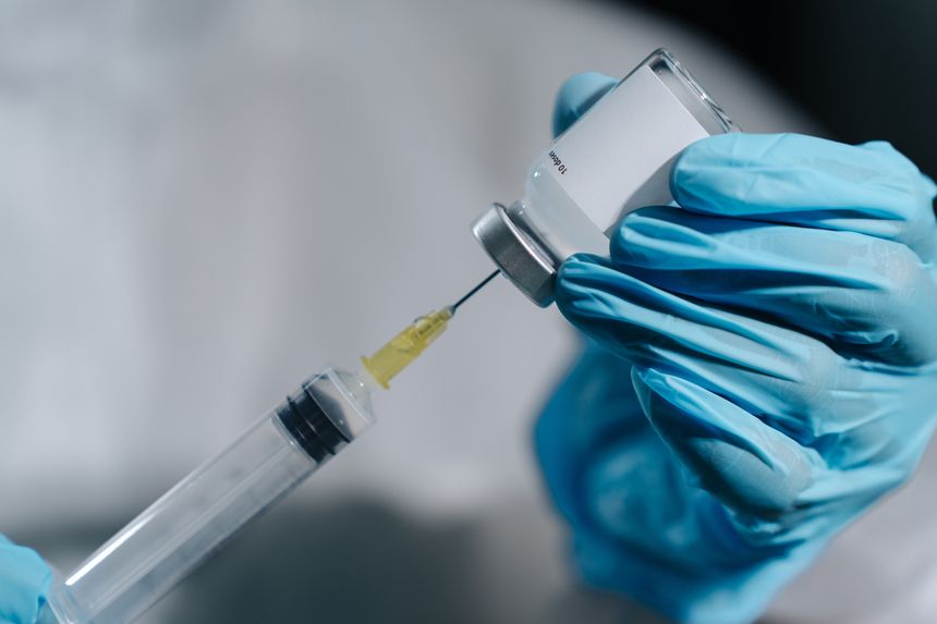 Extreme macro of syringe needle inserted into inverted vaccine vial with blank label, drawing Hantavirus antiviral drug, PPE gloves visible behind.