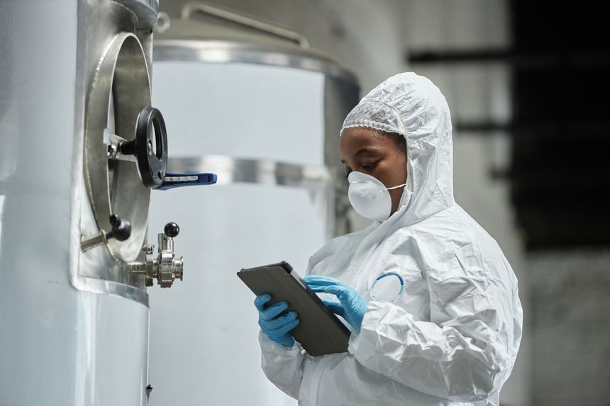 Medium shot of female African American technician in mask and hazmat suit using tablet computer inspecting stainless tanks while controlling fermentation process at cider factory, copy space