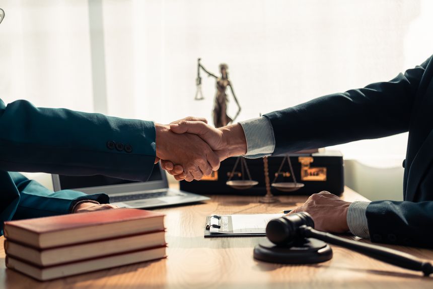 Close-up of lawyer and client shaking hands on table, symbolizing trust in legal contract with a hammer and a statue on the table, symbolizing justice.