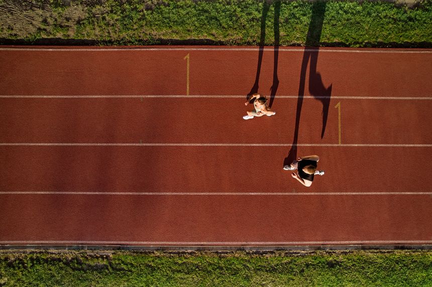 Drone top view of athletic man and woman jogging side by side on a red track near grass. Long shadows, evening sunlight. Concept of sport, endurance and healthy lifestyle.