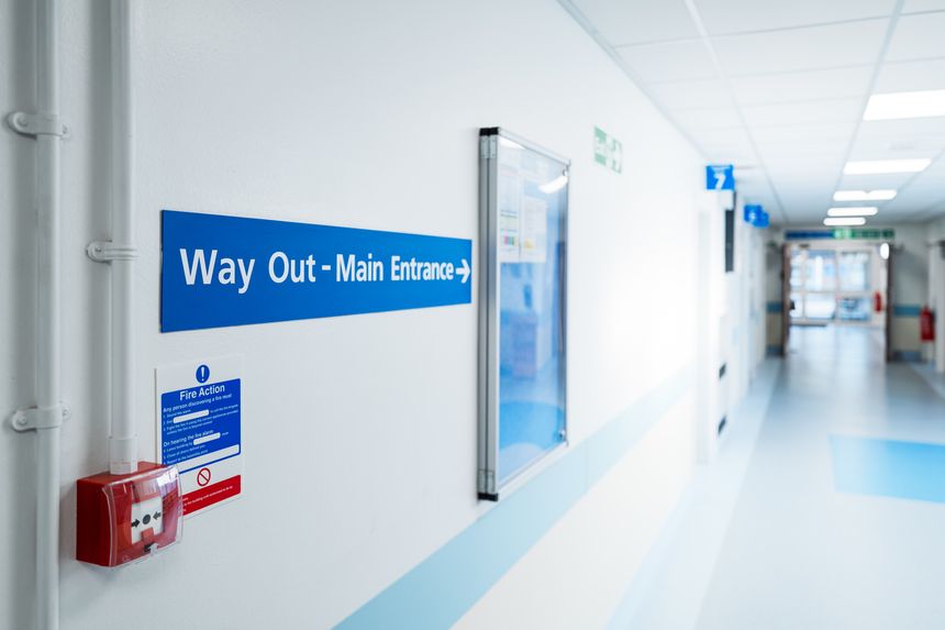 Shallow focus of a Way Out and Main Entrance sign seen within a typical NHS hospital in Britain. The corridor extends to outpatients and the distance double doors. A red alarm activator can be seen.
