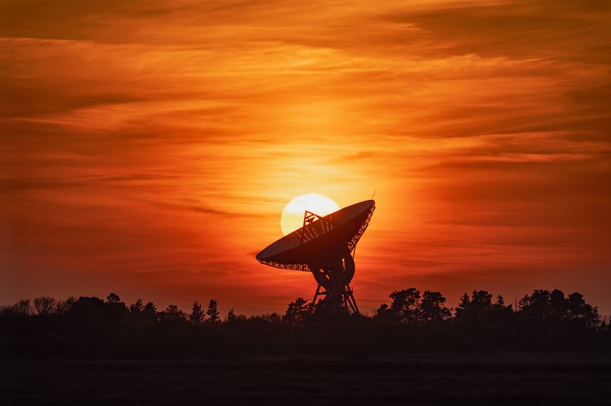 A large satellite dish with the sun stands in silhouette against a dramatic orange and red sunset sky. The structure, framed by trees and set against richly colored cloud formations, captures the contrast between technology and nature.