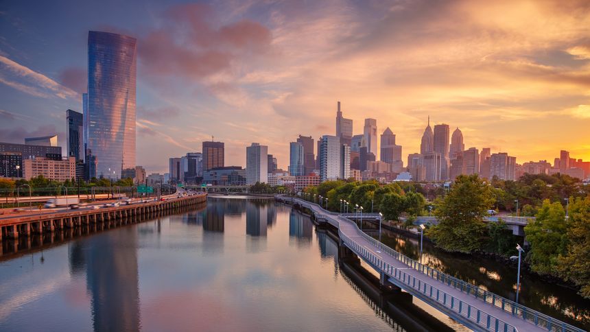 Philadelphia's skyline is seen at sunrise, featuring modern skyscrapers reflecting in the calm waters of the Schuylkill River. A curved pedestrian pathway follows the riverbank, surrounded by lush greenery transitioning into autumn colors. The sky is a blend of warm oranges and yellows, enhancing the serene morning atmosphere. Notable buildings, including the tall curved one on the left, dominate the landscape, highlighting the city's architectural diversity. The scene captures the tranquil beauty of the city at dawn.