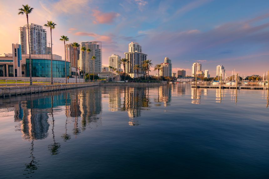 Skyscrapers line the St. Petersburg, Florida waterfront, their reflections shimmering on the calm water. The skyline features modern high-rises and palm trees, set against a colorful sunset with pink and orange hues. A sleek building with glass facades stands to the left, and several boats are moored along the docks on the right. The scene is serene, highlighting the city's urban beauty and scenic harbor during dusk.