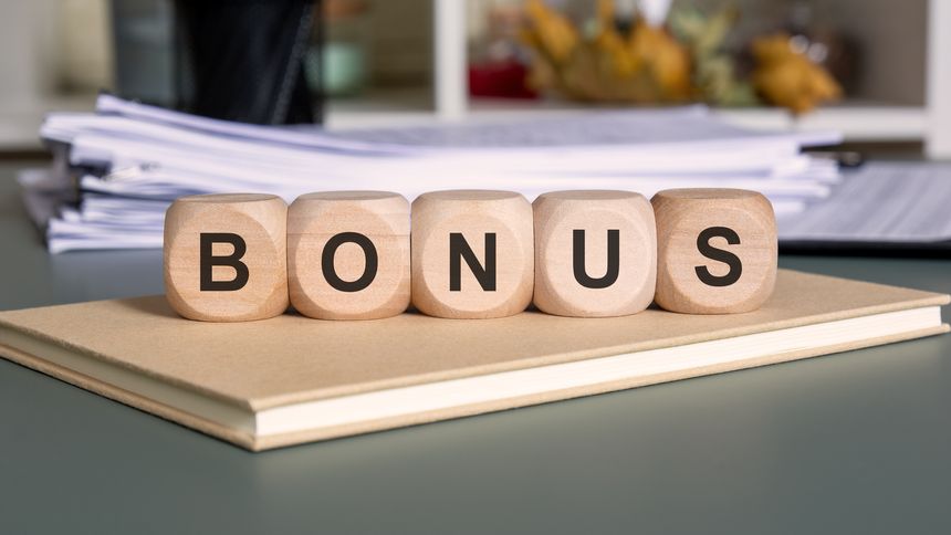 wooden blocks arranged to form the word BONUS are placed on a closed notebook with a pile of documents in the background. The image conveys themes of performance reward, employee incentive, and business success.