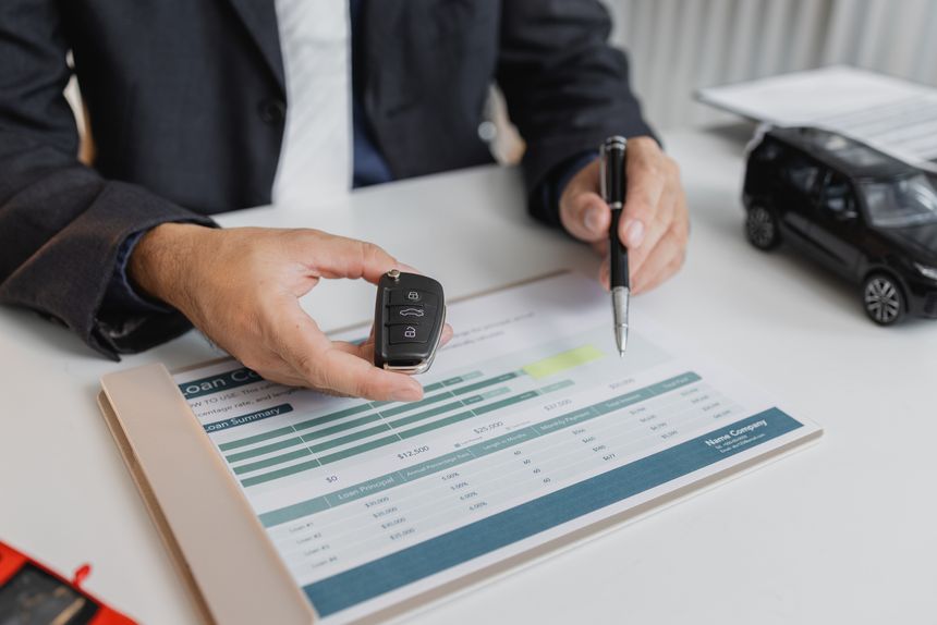 A businessman in a suit is calculating car loan options using a calculator and comparison chart, with model cars and car keys on the desk, symbolizing finance, leasing, and investment planning.