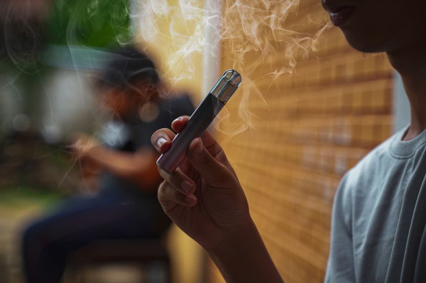 Asian male teenager holds a smoked e-cigarette in his hand while sitting during recess in a hidden corner with his friends at school.