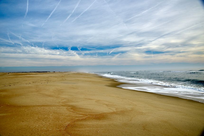 The scene shows a large sandy beach in Assateague Island National Seashore. Gentle waves from the ocean roll onto the shore, leaving marks in the sand. The sky is covered with scattered clouds, and there are traces of airplane contrails adding interest to the horizon. The beach stretches far into the distance, offering open space and a sense of emptiness.