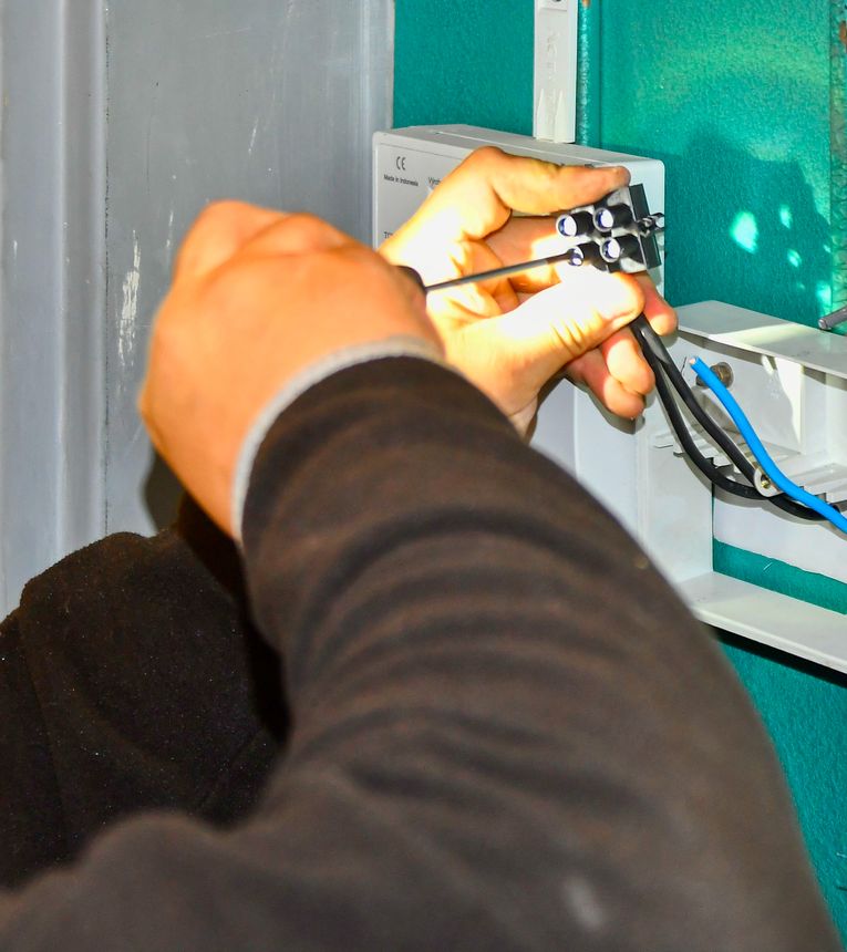 A person's hands are shown connecting electrical wires to a terminal block within a white junction box, highlighting a precise installation process