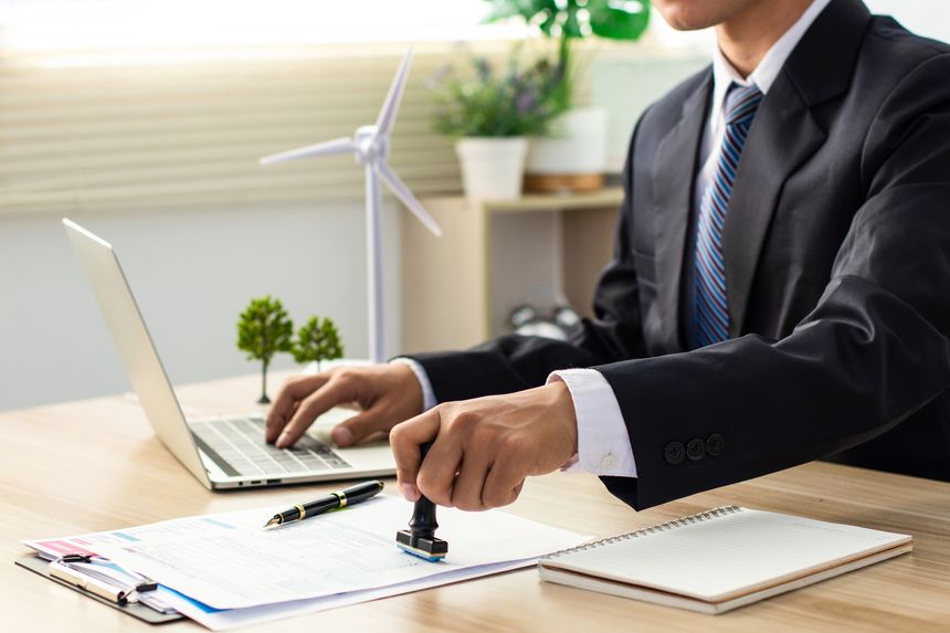 A businessman in a suit stamps his approval on a document, with a miniature wind turbine visible on the desk, indicating a focus on renewable energy in business. ESG. Co2. Net zero. Green energy. CSR.