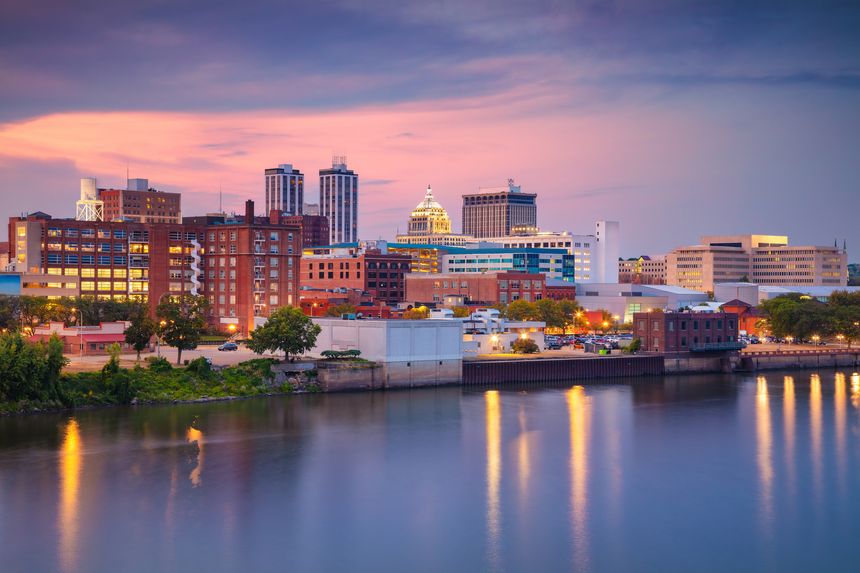 Peoria, Illinois during twilight blue hour features a skyline with illuminated buildings reflecting off a calm river. Prominent structures, including a white dome, stand out against the dusk sky with hues of purple and pink. Trees and greenery line the riverbank, and the city's architecture includes a mix of modern and classic designs. The overall ambiance is serene and picturesque, capturing the transitional light during early evening.