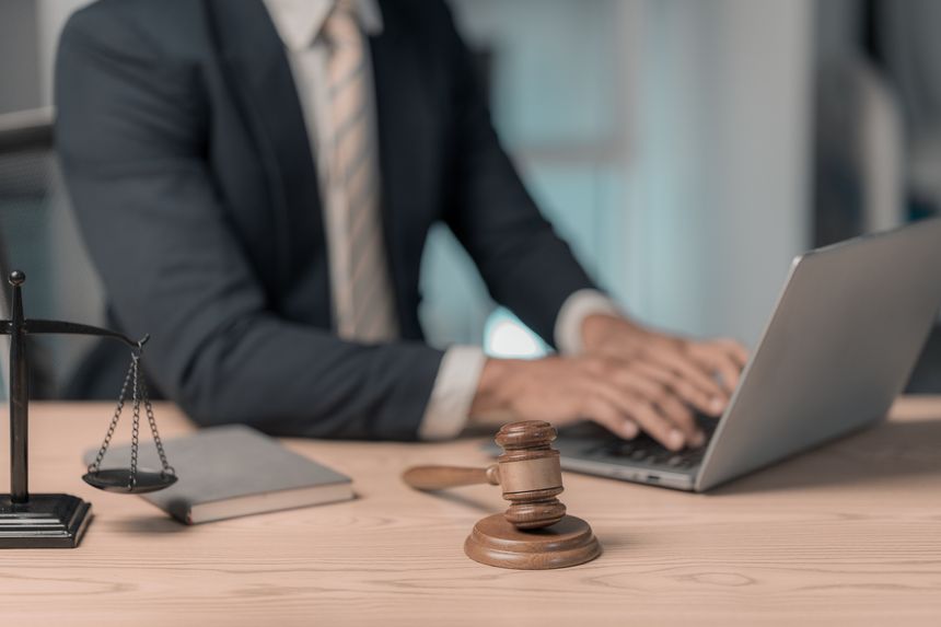 A lawyer or legal professional sits at a desk. On the desk are several objects related to law and a laptop, a clipboard with documents, a hammer, and a statue of the Goddess of Justice.