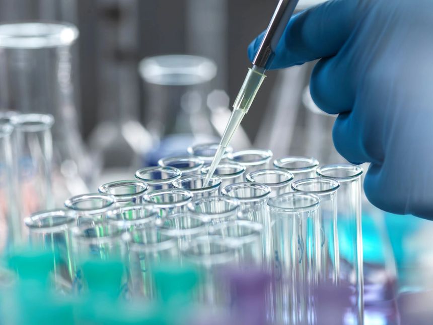 A gloved hand carefully uses a pipette to dispense liquid into a row of laboratory test tubes. The scene highlights scientific research.