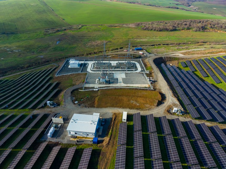 A solar energy farm shows rows of solar panels adjacent to a control building in a green countryside area during daylight hours.