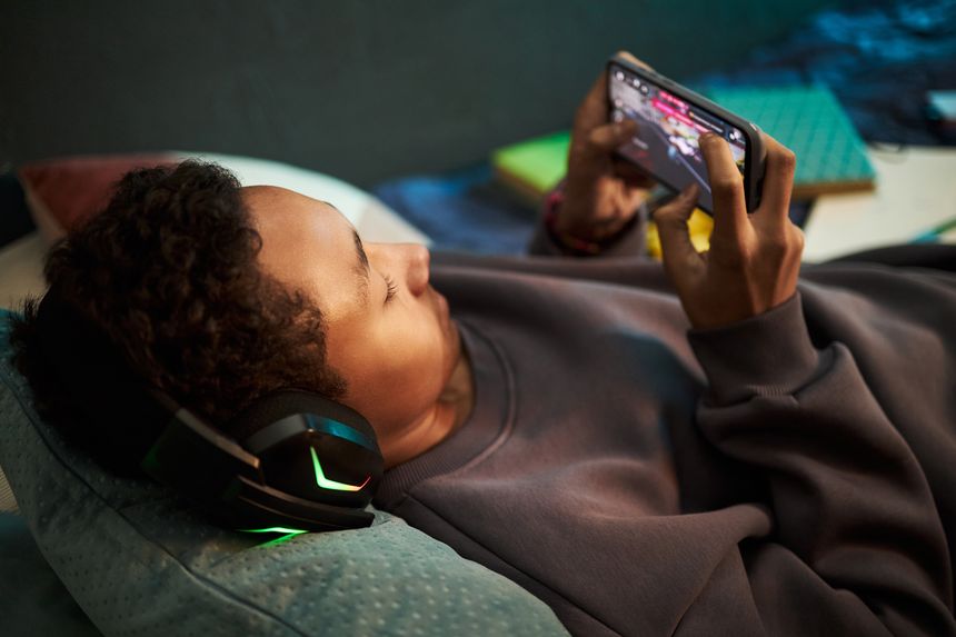 Black teenager boy lying on bed wearing headphones, playing mobile game on smartphone holding device with both hands focused on screen relaxing in casual home environment