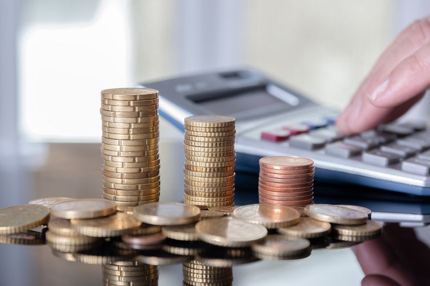 Coins stacked in a pile on a table and hand typing on a calculator forming part of a blurred background.