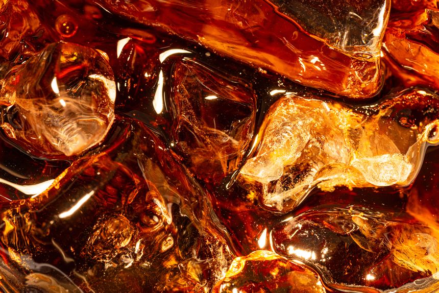High-angle macro shot of sparkling brown soda or cola with crystal clear ice cubes. The image captures the refreshing texture, golden light reflections, and carbonated bubbles, perfect for summer drinks or beverage concepts.