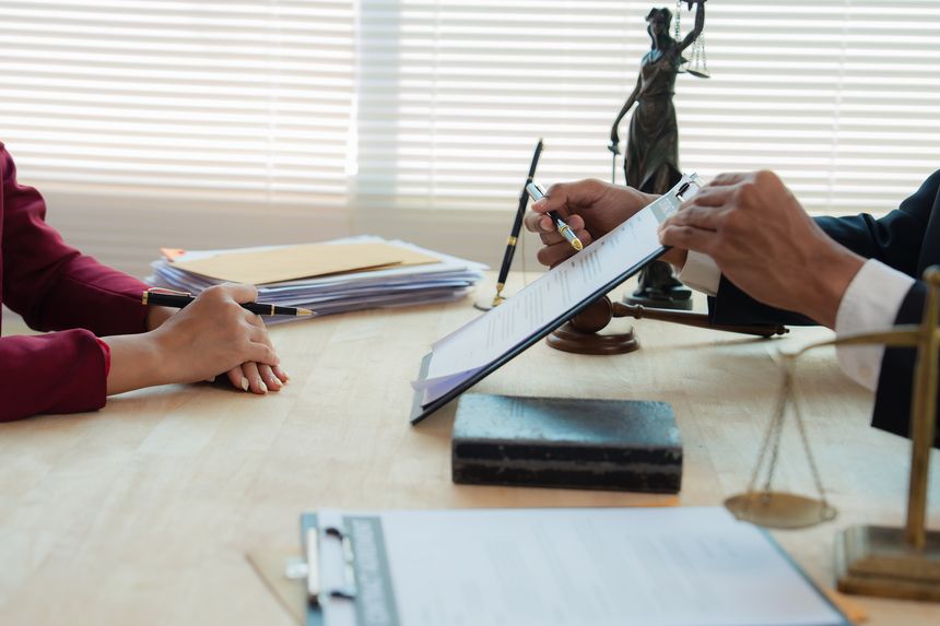 Close up of justice scales and judge gavel on wooden desk with cash bundle and lawyer consulting client about financial lawsuit in blurred background.