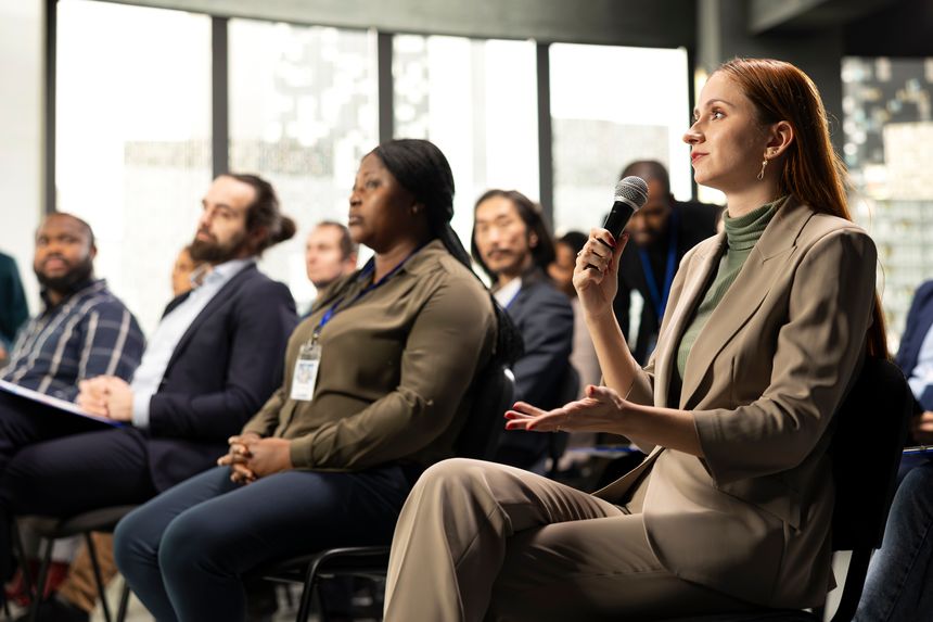 Young adult businesswoman in audience interacting with presenter, asking questions and engaging in debate. Mixed age participants include managers, employees and investors at summit.