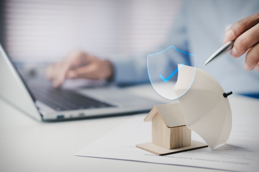Modern home insurance protection concept, wooden house model under umbrella beside laptop, digital security shield overlay, clean corporate workspace representing property coverage, financial safety