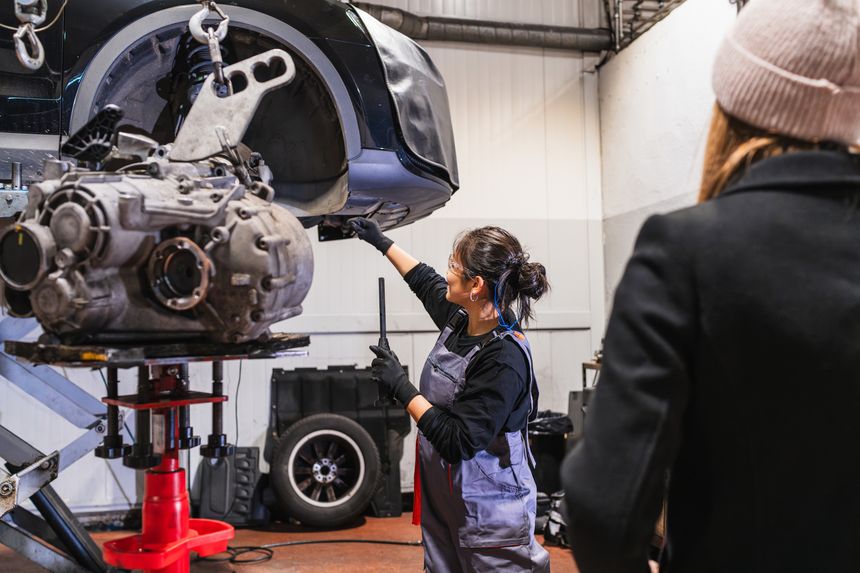 Female mechanic is installing a gearbox in a car at a repair shop, while a customer observes her work