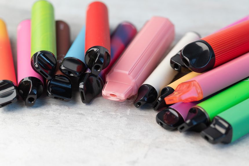 Collection of various colorful disposable e-cigarettes and vape pens scattered on a light grey background.