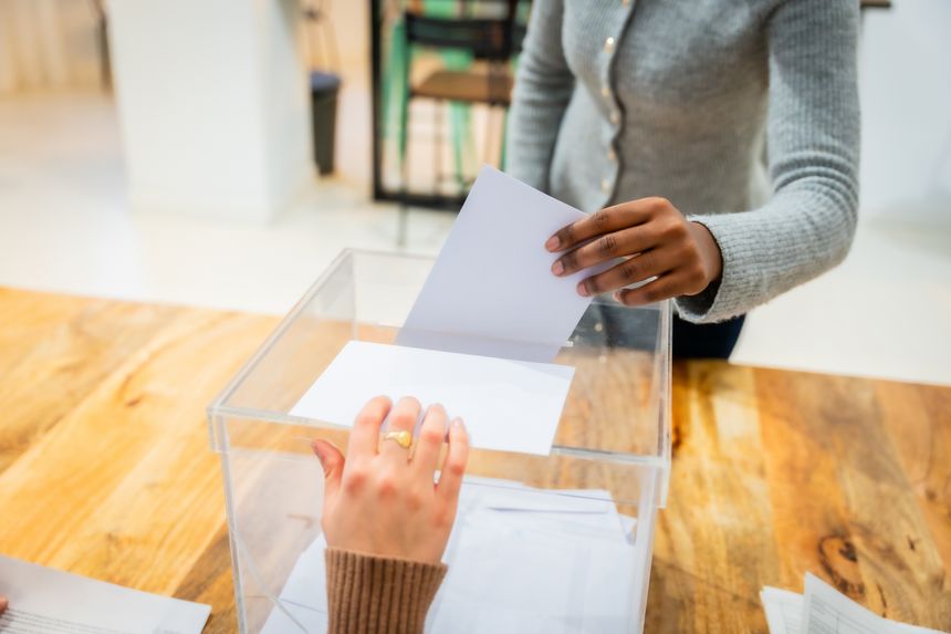 Two people are casting ballots into a clear ballot box, participating in an election process and exercising their right to vote for democratic representation