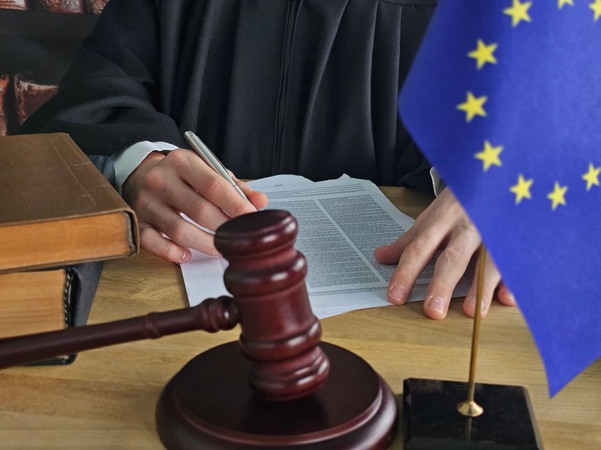 Courtroom scene shows judge writing on legal documents with an EU flag and gavel in view during a formal session in a judicial setting