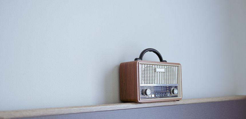 A vintage-inspired portable radio with a classic wood grain finish and analog tuning dial sits elegantly on a wooden shelf against a clean, minimalist light-colored wall. The image captures a nostalgic, cozy atmosphere perfect for home decor and lifestyle concepts.