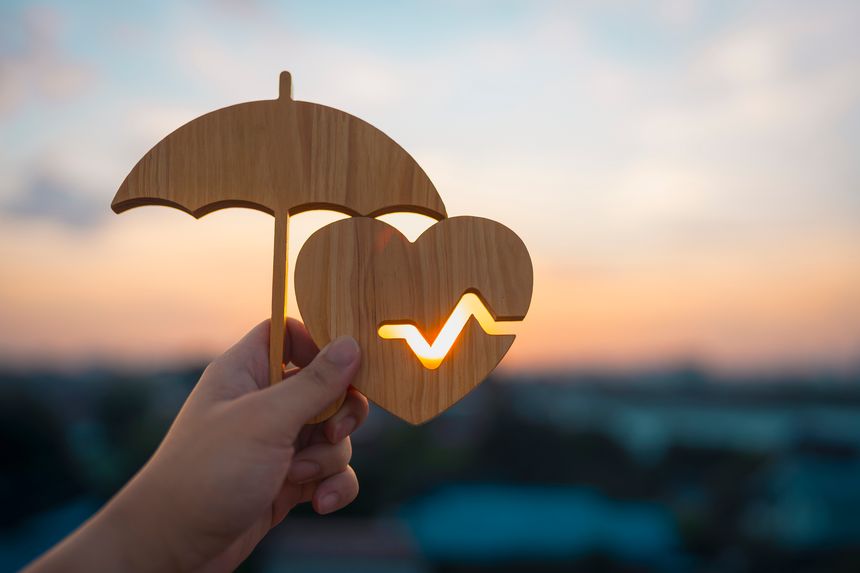 Health insurance and life protection concept with hand holding wooden umbrella over heart with pulse line silhouette against sunset background, medical safety and wellness security.