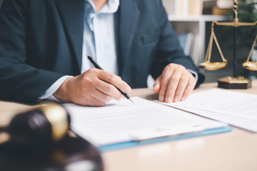 Lawyer in suit signing a contract on clipboard at office desk with justice scales in background, representing legal services, compliance review, agreement approval and documentation.