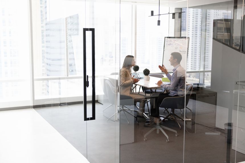 Two male and female business leaders discussing company management strategy in meeting room with glass walls, sitting at negotiation table, talking. Wide shot with copy space