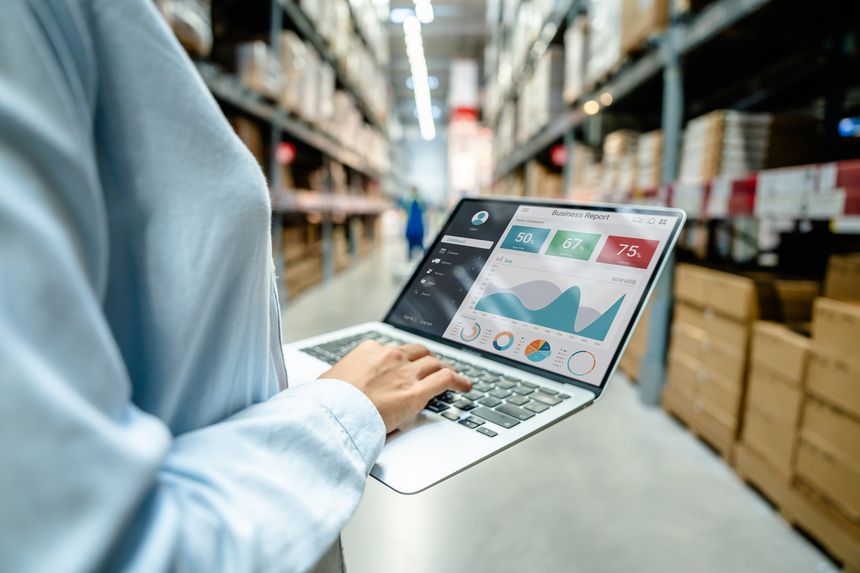 Employee using a laptop with business analytics dashboard in a modern warehouse, showing real-time data monitoring and smart supply chain management for logistics and inventory control.