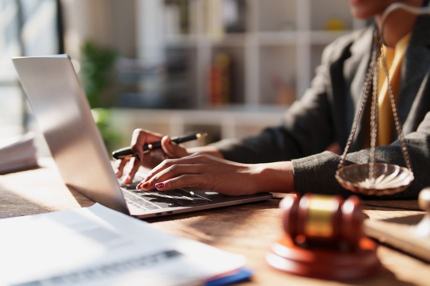 Young lawyer typing on a laptop in an office, offering legal advice with scales of justice and a gavel on the desk, close-up on hands and documents